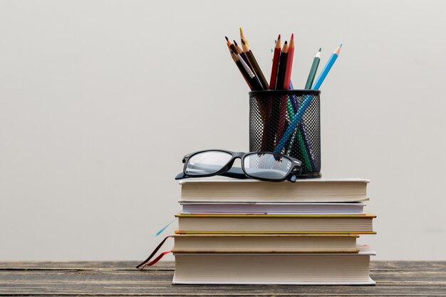 Concepto de regreso a la escuela con gafas, libros, lápices en soporte en vista lateral de madera y pared blanca.