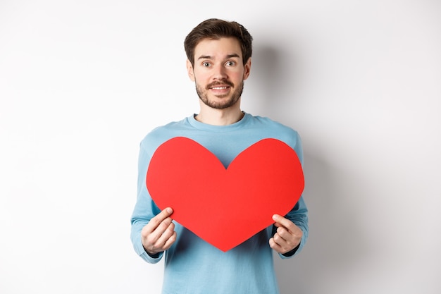 Concepto de relación y amor. Hombre caucásico hermoso en suéter que sostiene el recorte rojo grande del corazón del día de tarjetas del día de San Valentín y que sonríe, que confiesa en la fecha, que se coloca en el fondo blanco.