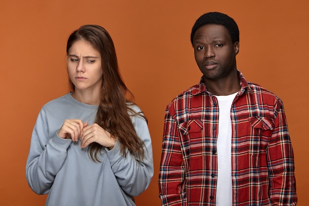 Foto gratuita concepto de relaciones y emociones humanas negativas. molesta mujer blanca joven vestida de manera informal de pie en postura cerrada, con mirada preocupada, asustada de su abusador marido de piel oscura