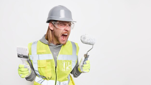 Concepto de reparación y renovación. Constructor masculino irritado molesto exclama en voz alta sostiene la brocha y el rodillo