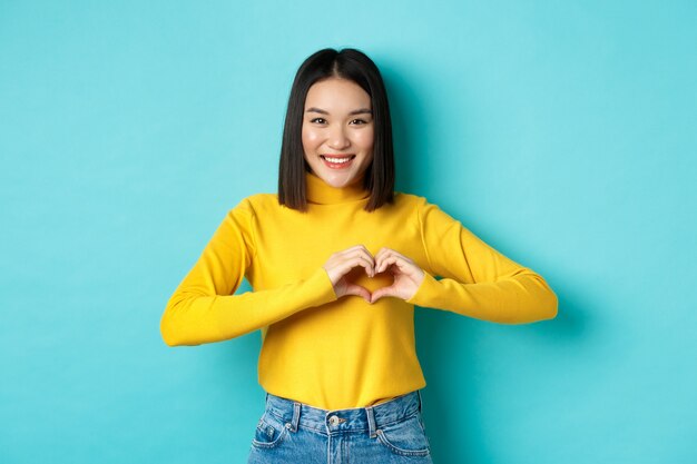 Concepto de romance y día de San Valentín. Hermosa mujer asiática muestra te amo, gesto de corazón y sonriente, de pie contra el fondo azul.