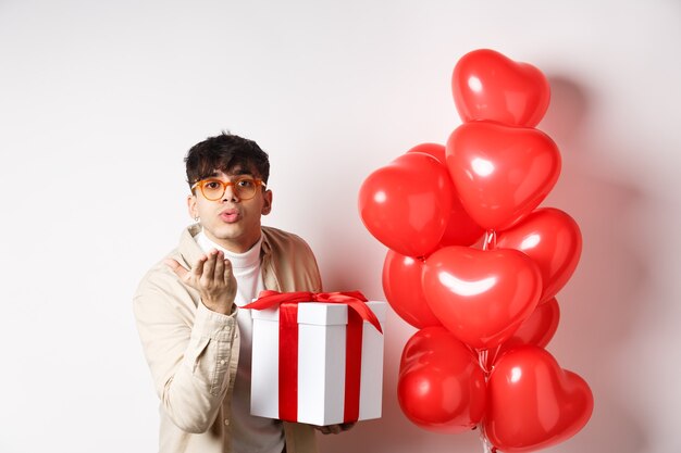 Concepto de romance y día de San Valentín. Hombre moderno romántico con regalo especial para amante y enviando beso de aire a la cámara, de pie cerca de globos de corazones, fondo blanco.