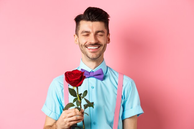 Concepto de romance y día de San Valentín. Hombre romántico con rosa roja en cita con su amante, de pie en elegante pajarita sobre fondo rosa.