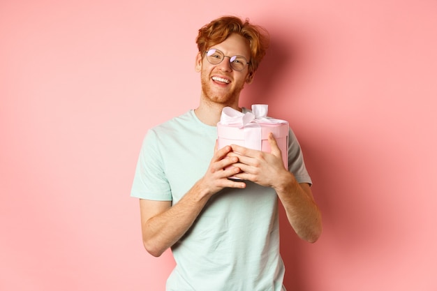 Concepto de romance y día de San Valentín. Novio pelirrojo feliz recibe regalo romántico, caja de abrazos con presente y diciendo gracias, sonriendo agradecido, fondo rosa.