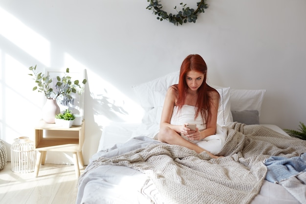 Concepto de ropa de cama, ocio y tecnologías modernas. Toma interior de una hermosa joven pelirroja que se queda en la cama durante el día, navega por internet en el teléfono celular, envía mensajes a amigos a través de las redes sociales