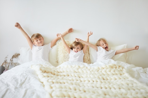 Concepto de ropa de cama, sueño, descanso y relajación. Toma en interiores de tres niños que se sienten somnolientos mientras se despiertan temprano en la mañana antes de la escuela. Dos hermanos y una hermana bostezando y estirando en la cama