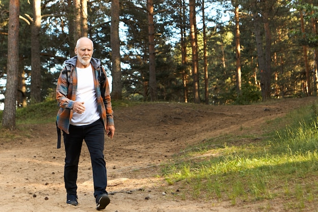 Concepto de salud, actividad, bienestar, edad y personas. Hombre europeo barbudo activo autodeterminado de unos sesenta años caminando rápido mientras camina en el bosque de montaña, con mirada centrada en confianza