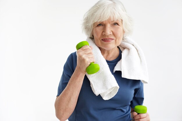 Concepto de salud, bienestar, actividad, madurez y edad. Anciana optimista en entrenamiento de jubilación en el interior con una toalla blanca alrededor del cuello, haciendo ejercicio con un par de pesas verdes y sonriendo