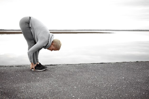 Concepto de salud, bienestar y actividad. Tiro de longitud completa de mujer de pelo corto en forma en zapatillas y ropa deportiva haciendo ejercicios físicos al aire libre, posando en el lago en pose de yoga de inclinación hacia adelante