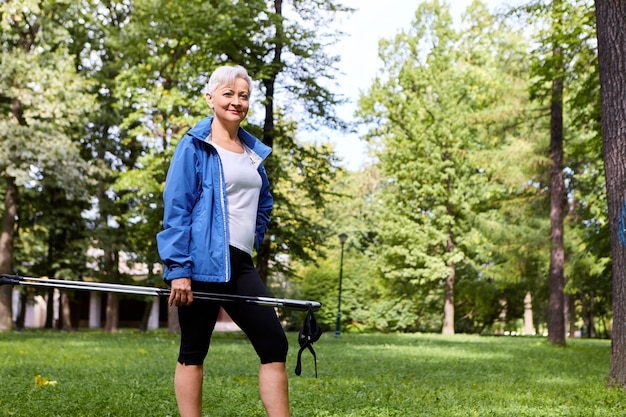 Concepto de salud, bienestar, vitalidad, recreación y actividad Vista de verano al aire libre de una elegante y segura mujer de sesenta años posando contra pinos, sosteniendo un bastón nórdico y sonriendo