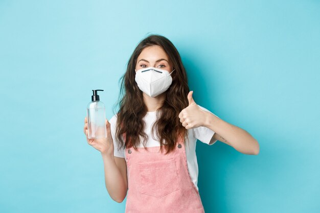 Concepto de salud, coronavirus y distanciamiento social. Mujer joven con mascarilla, respirador y mostrando desinfectante de manos con el pulgar hacia arriba, recomendando un fondo azul antiséptico.