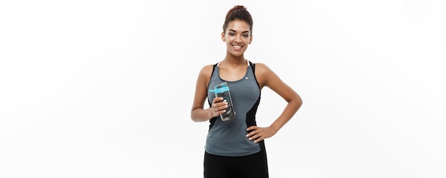 Concepto de salud y fitness hermosa chica afroamericana en ropa deportiva sosteniendo una botella de agua después del entrenamiento aislado sobre fondo blanco de estudio