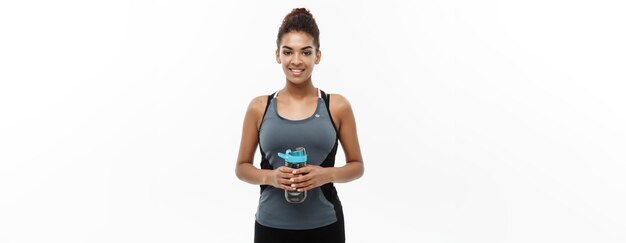 Concepto de salud y fitness hermosa chica afroamericana en ropa deportiva sosteniendo una botella de agua después del entrenamiento aislado sobre fondo blanco de estudio