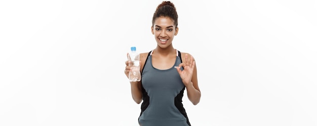 Concepto de salud y fitness hermosa chica afroamericana en ropa deportiva sosteniendo una botella de agua de plástico después del entrenamiento aislado sobre fondo blanco de estudio