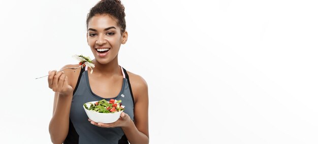 Concepto de salud y fitness Hermosa dama africana americana en ropa de fitness en dieta comiendo ensalada fresca Aislada sobre fondo blanco
