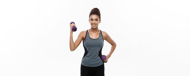 Concepto de salud y fitness Hermosa dama africana americana en ropa de fitness entrenamiento con mancuernas aislado sobre fondo blanco.