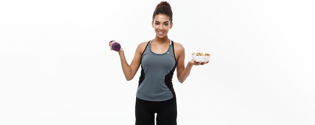 Concepto de salud y fitness Hermoso deportivo afroamericano en dieta con mancuerna y ensalada fresca en las manos Aislado sobre fondo blanco de estudio