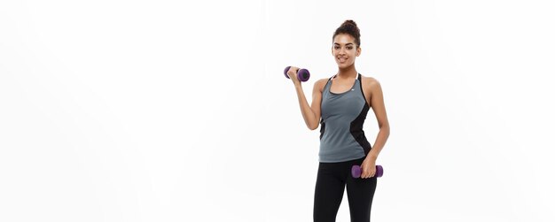 Concepto de salud y fitness Longitud total de retrato Hermosa dama africana americana en ropa de fitness gris entrenamiento con mancuernas Aislado sobre fondo blanco