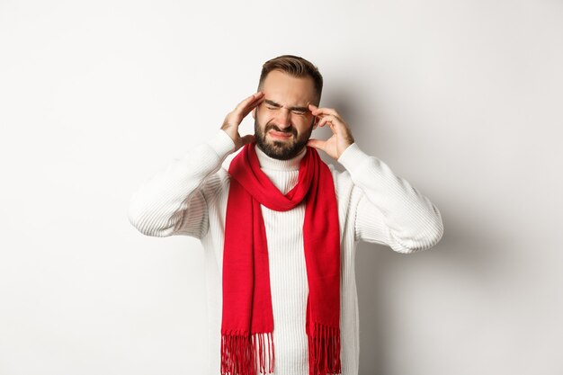 Concepto de salud. Hombre con dolor de cabeza severo, tocarse la cabeza y hacer muecas de migraña, de pie en suéter de invierno y pañuelo rojo, fondo blanco.