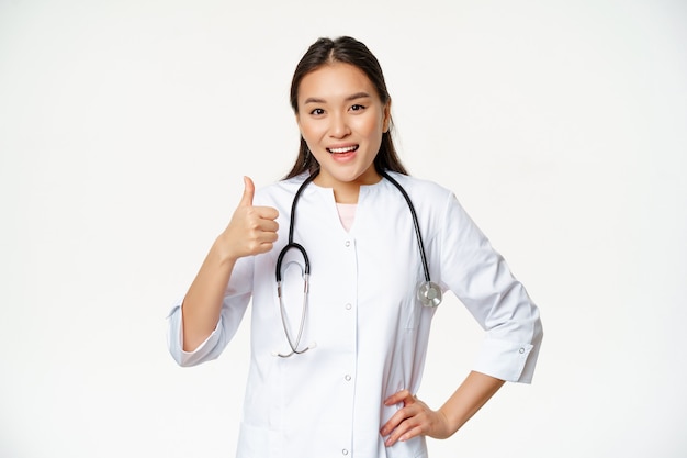 Concepto de salud y hospital. Sonriente médico asiático muestra los pulgares hacia arriba en señal de aprobación, dice que sí, luciendo satisfecho, de pie en uniforme sobre fondo blanco.