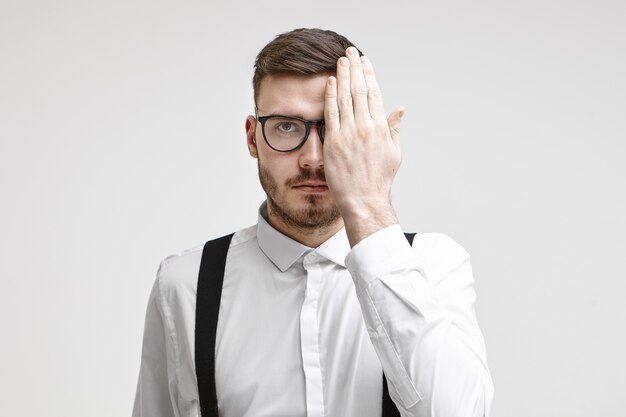 Concepto de salud, medicina, personas, vista, óptica, gafas y lentes de contacto. Hombre joven serio con barba que cubre un ojo mientras se le examinan los ojos durante el examen de la vista ocular