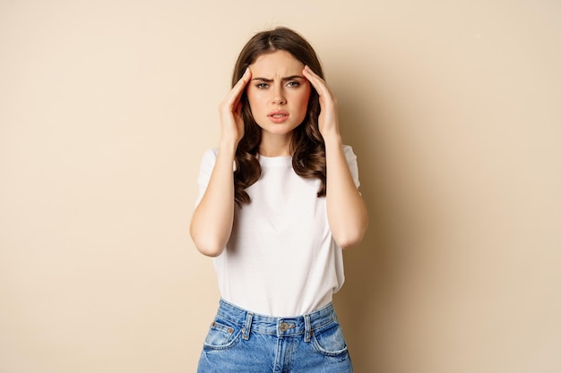 Concepto de salud y mujer. Retrato de niña sintiéndose enferma, tocando las sienes de la cabeza, sintiendo dolor de cabeza, migraña, de pie sobre fondo beige