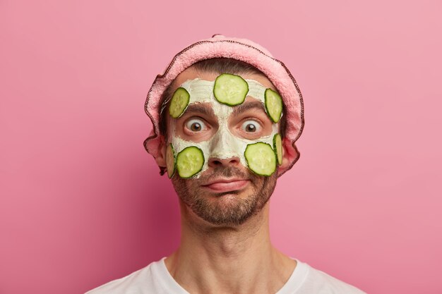 Concepto de salud de la piel. Hombre emotivo aturdido con los ojos bien abiertos, tiene tratamientos de cosmetología en casa, limpia la tez con mascarilla nutritiva y pepino