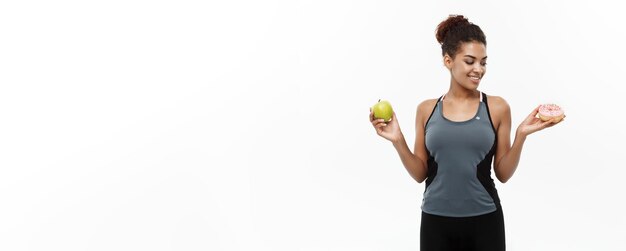 Concepto saludable y dietético Hermoso afroamericano deportivo toma una decisión entre donut y manzana verde Aislado sobre fondo blanco
