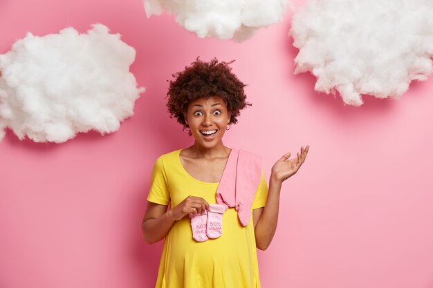 Concepto saludable de maternidad y embarazo. La futura mamá alegre y joven mira con alegría y gesticula con vacilación, anticipa a la niña, compra calcetines y un mono rosados, expresa felicidad