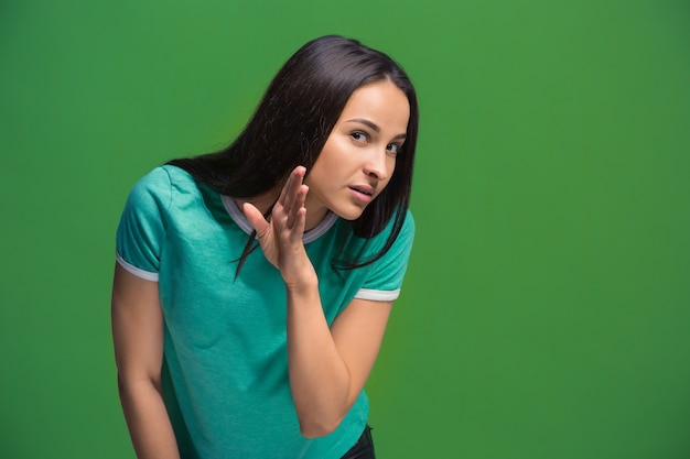 Concepto secreto de chismes. Mujer joven susurrando un secreto detrás de su mano. Mujer de negocios aislada sobre fondo de moda estudio verde. Joven mujer emocional. Las emociones humanas, el concepto de expresión facial.