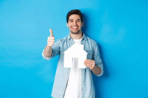 Concepto de seguros, hipotecas y bienes raíces. Cliente satisfecho mostrando el modelo de la casa y el pulgar hacia arriba, sonriendo complacido, de pie contra el fondo azul.