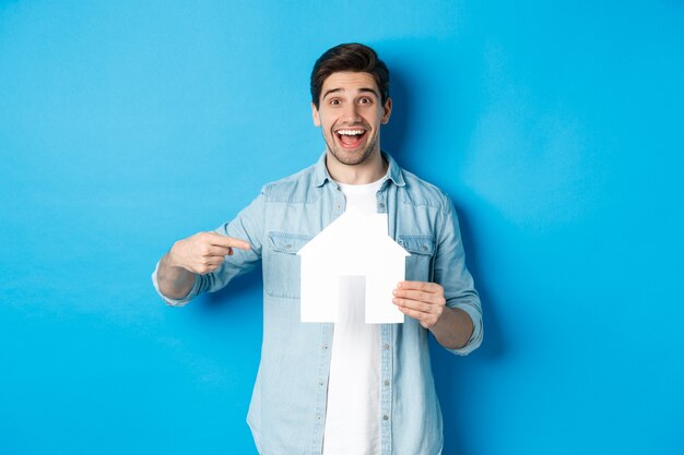 Concepto de seguros, hipotecas y bienes raíces. Hombre sorprendido apuntando al modelo de la casa y sonriendo, buscando apartamento para alquilar o comprar, de pie contra el fondo azul.