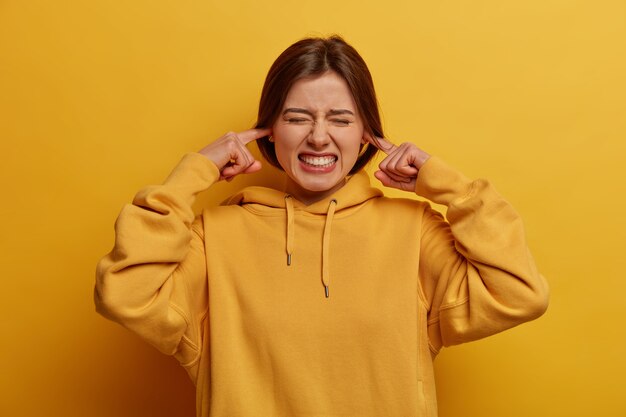 Concepto de sentimientos negativos. Mujer irritada tapona los oídos y aprieta los dientes, se queja del ruido, ignora las conversaciones desagradables, usa una sudadera con capucha informal