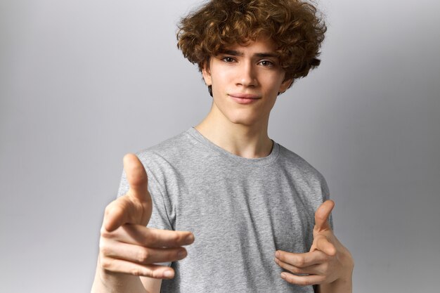 Concepto de signos, gestos y símbolos. Retrato de un joven guapo con cabello ondulado y ojos marrones apuntando con el dedo índice a la cámara, desafiándote o eligiéndote, mirando con una sonrisa segura