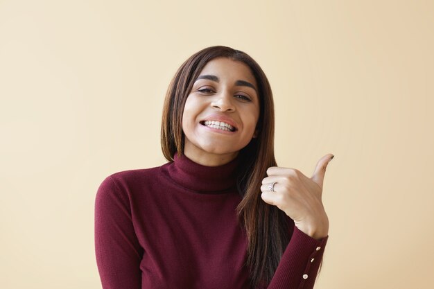 Concepto de signos, símbolos y gestos. Alegre joven dama de raza mixta con cabello largo y liso sonriendo ampliamente y mostrando los pulgares hacia arriba gesto como señal de aprobación, respeto y agrado