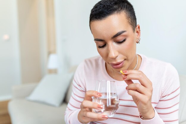 Concepto de sufrimiento de un dolor terrible Hermosa mujer triste, triste, infeliz, con problemas, débil, con el pelo negro y ropa informal, está sentada en un sofá y tomando una pastilla