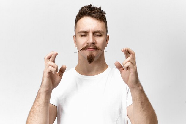 Concepto de superstición y expectativa. Imagen de un guapo joven supersticioso hipster barbudo cerrando los ojos y cruzando los dedos para tener buena suerte, esperando que todos sus sueños se hagan realidad