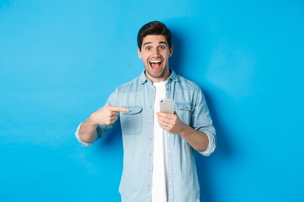 Concepto de tecnología, aplicaciones y compras online. Feliz el hombre sonriente señalando con el dedo al teléfono, de pie asombrado contra el fondo azul.