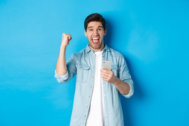 Concepto de tecnología, aplicaciones y compras online. Hombre emocionado gritando sí y haciendo gesto de bomba de puño después de ganar en el teléfono inteligente, de pie sobre fondo azul