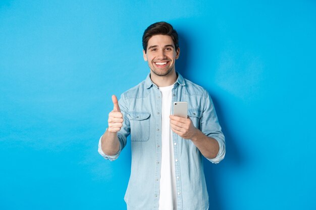 Concepto de tecnología, aplicaciones y compras online. Hombre satisfecho en ropa casual sonriendo, mostrando los pulgares hacia arriba después de usar la aplicación de teléfono inteligente, de pie sobre fondo azul