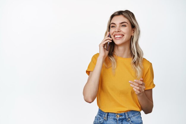 Concepto de tecnología celular Mujer joven sonriente que tiene una llamada telefónica hablando por teléfono móvil con expresión de cara feliz con camiseta amarilla y fondo blanco de jeans