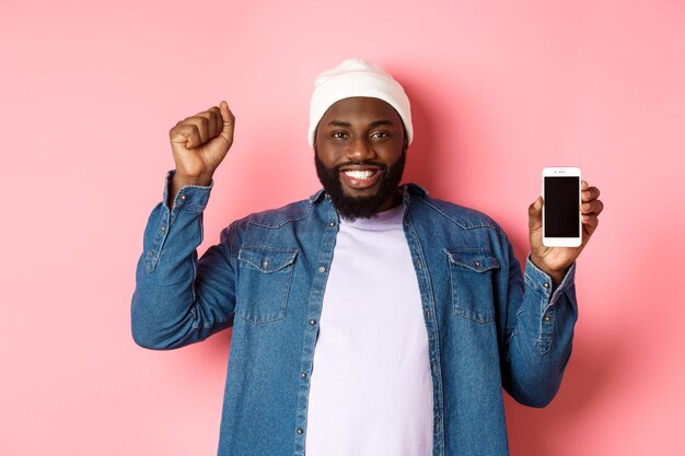 Concepto de tecnología y compras en línea. Alegre hombre negro regocijándose y mostrando la pantalla del móvil, levantando la mano satisfecho, triunfando mientras está parado sobre fondo rosa