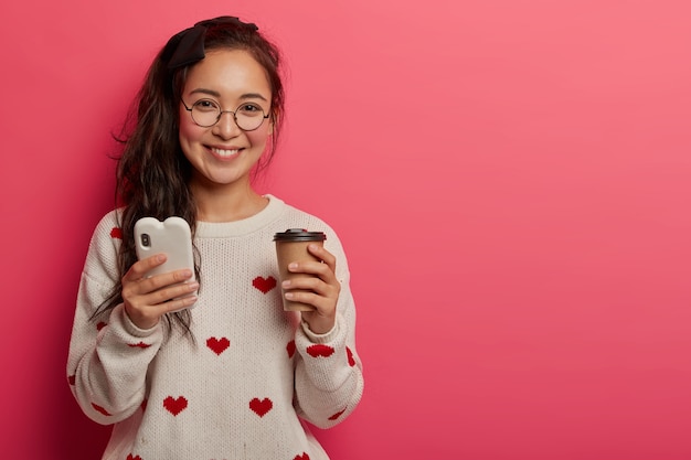 Foto gratuita concepto de tecnología, comunicación y estilo de vida. bastante joven con gafas redondas usa un teléfono inteligente para leer blogs y mensajes geniales, bebidas, café para llevar, aplicaciones de descargas, se encuentra en el interior.