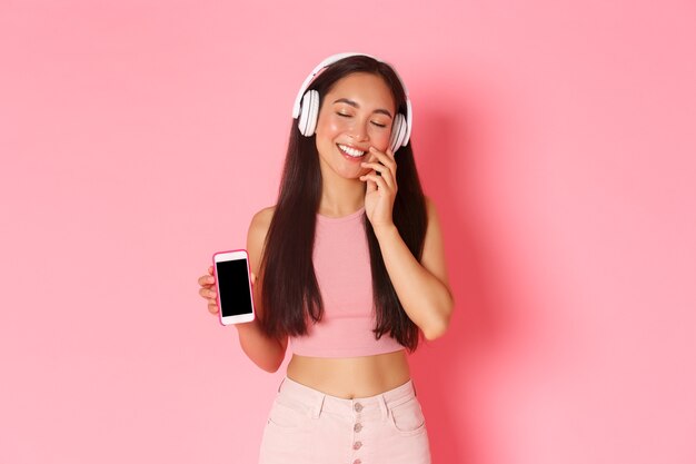 Concepto de tecnología, comunicación y estilo de vida en línea. Chica asiática encantadora y soñadora en auriculares, cerrar los ojos y tocar la mejilla tontamente, soñar despierto mientras escucha música, mostrando la pantalla del móvil