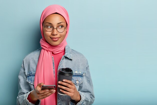 Concepto de tecnología y comunicación. Foto de mujer musulmana complacida con velo rosa, usa nueva aplicación de teléfono inteligente instalada, sostiene café para llevar, usa anteojos redondos, se para en el interior sobre una pared azul