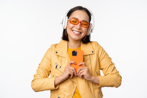 Concepto de tecnología Elegante chica asiática en auriculares con smartphone bailando y cantando escuchando música de pie sobre fondo blanco.
