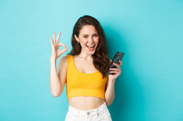 Concepto de tecnología y estilo de vida. La mujer atractiva confiada dice que sí, mostrando un signo bien a algo en línea, sosteniendo un teléfono inteligente, de pie sobre un fondo azul.
