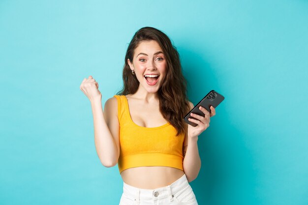 Concepto de tecnología y estilo de vida. Mujer emocionada ganando en línea, sosteniendo un teléfono inteligente y haciendo un gesto de bomba de puño sí, celebrando, lograr el objetivo en la aplicación, fondo azul.
