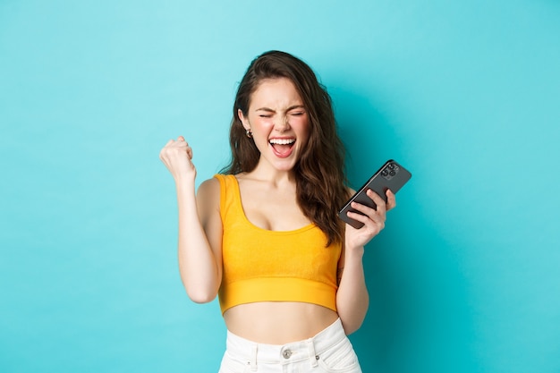 Concepto de tecnología y estilo de vida. La mujer satisfecha logra el éxito, gana en el teléfono móvil, levanta el puño y grita que sí con una expresión alegre, de pie sobre un fondo azul.