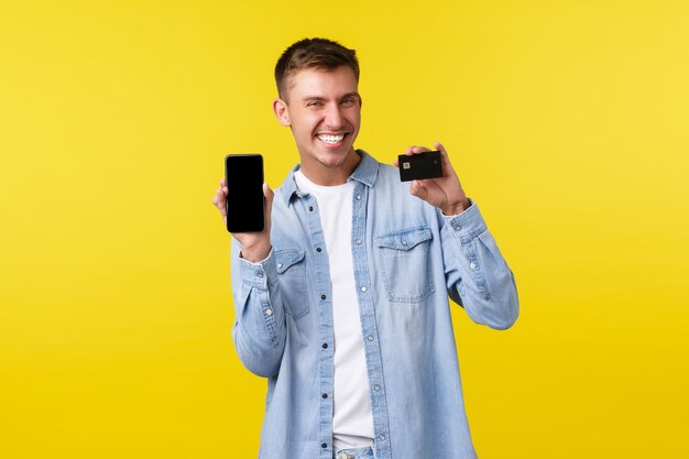 Concepto de tecnología, estilo de vida y publicidad. Chico rubio feliz contento que muestra la tarjeta de crédito y el teléfono móvil, recomendando la aplicación para ir de compras, fondo amarillo de pie. Copia espacio